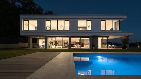 modernes großes Wohnhaus mit Flachdach und vielen Holzalufenstern bei Nacht. In Inneren des Hauses brennt Licht und im Vordergrund sieht man einen Pool.