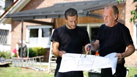 Zwei Männer stehen mit Bauplan vor einen Haus mit Wintergarten
