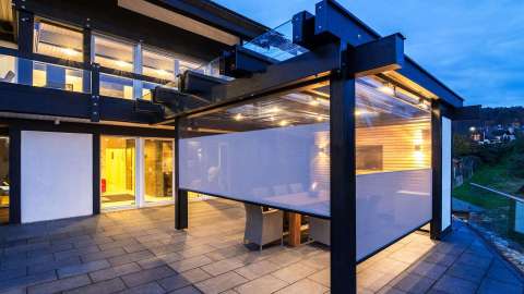 Terrasse mit Glashaus mit Textilscreens bei Dämmerung, drinnen beleuchtet