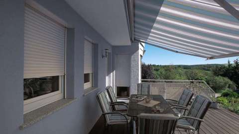 Terrasse an einem hellblauen Haus mit ausgefahrener Markise. Die Rollläden vor den Fenster von halb geschlossen und auf der Terrasse steht ein Tisch mit Stühlen
