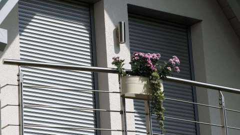 Balkongeländer mit Blumenkasten vor zwei bodentiefen Fenster mit runtergelassenem Rollo