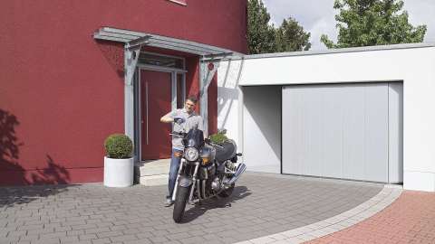 Seiten-Sektionaltor in einer Garage neben einem roten Haus. Davor steht ein Mann mit seinem Motorrad