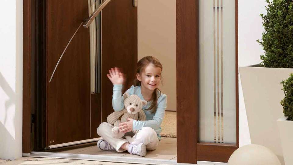 Kind mit Teddybär sitzt in einer geöffneten Kunststoff-Haustür und winkt